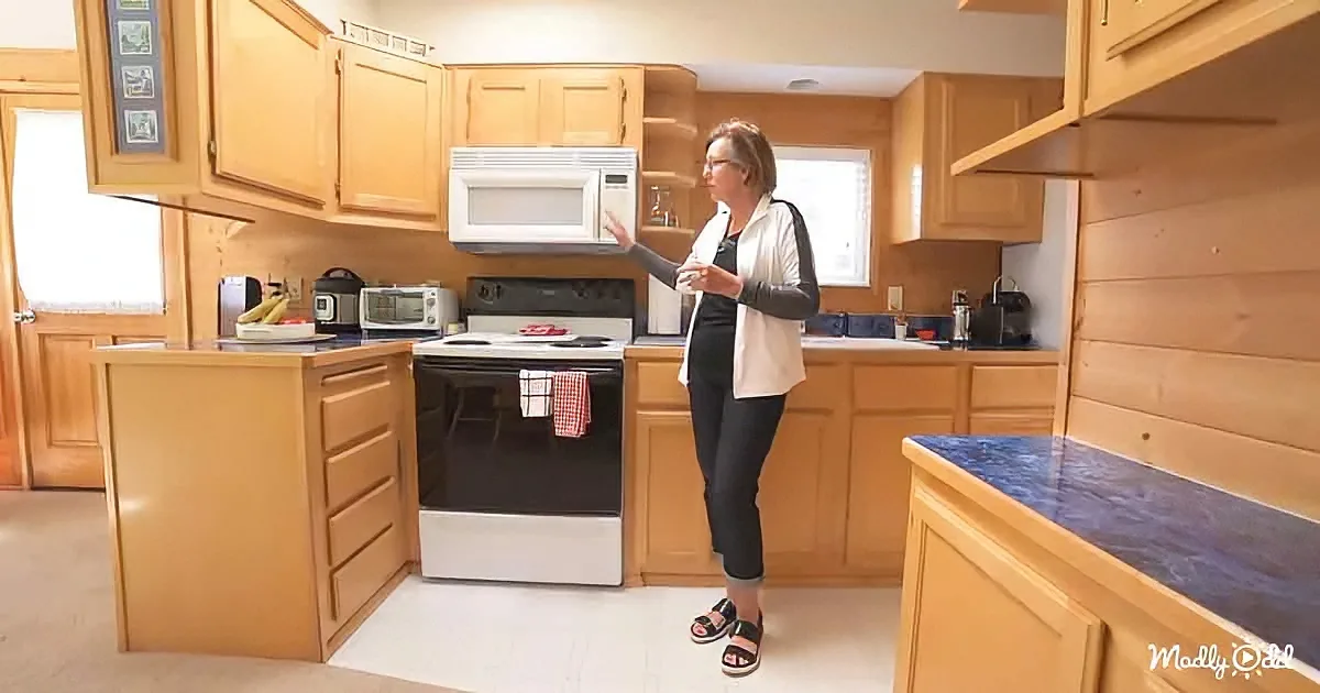 A woman stands in a kitchen, pointing at a microwave above a stove with a red and white checkered towel.
