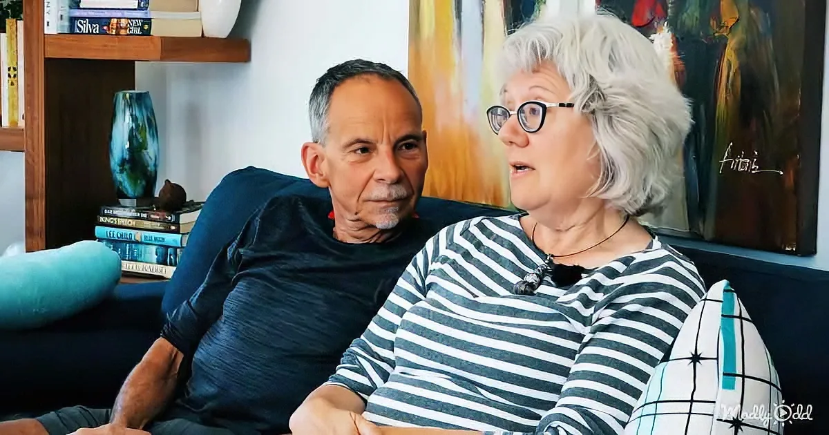 A man and woman sit on a couch, the woman wearing glasses and a striped shirt, in a cozy living room with books and art.
