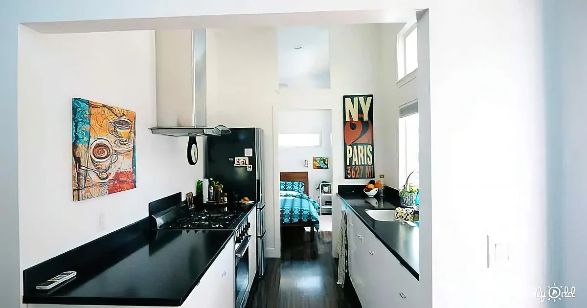 A modern kitchen with black countertops, white cabinets, and a view into a bedroom with a bed and artwork.
