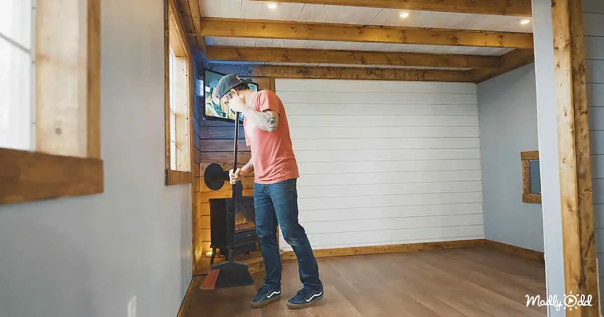 A man in a pink shirt and jeans cleans a fireplace with a broom in a rustic room with wooden beams and white walls.