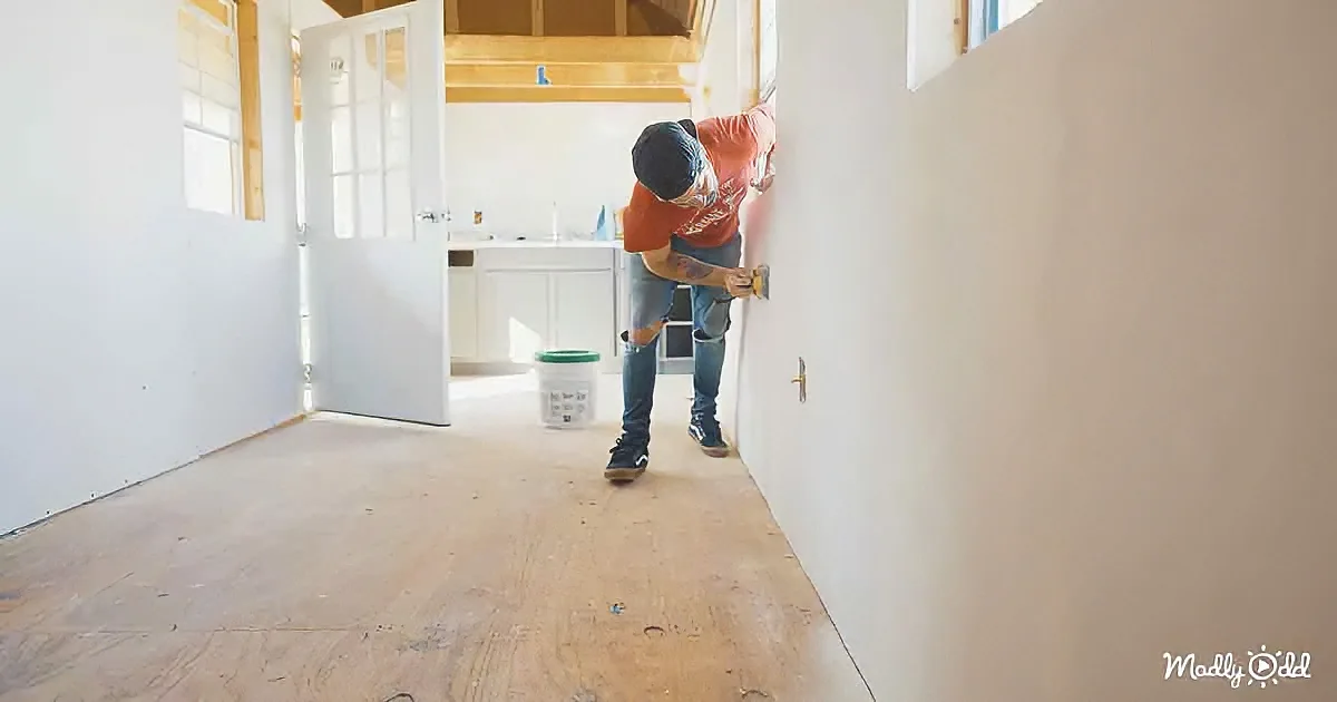 A man in a red shirt and jeans is painting a wall in a room with a wooden floor and white walls.