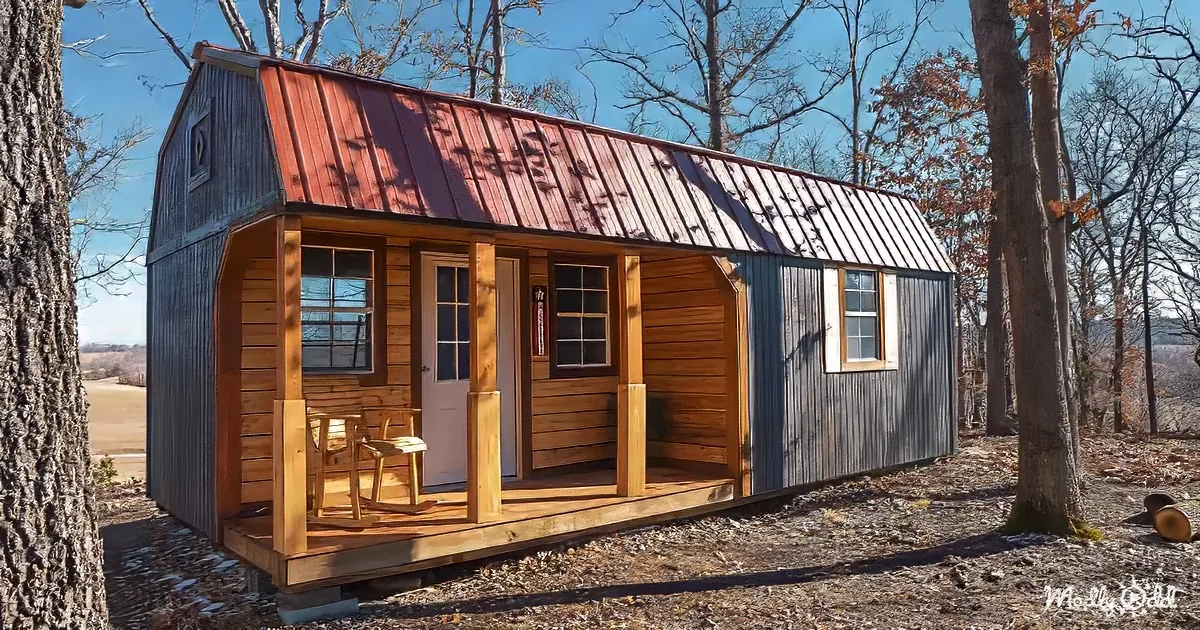 A rustic cabin with a red metal roof and wooden porch sits amidst bare trees and a clear blue sky.