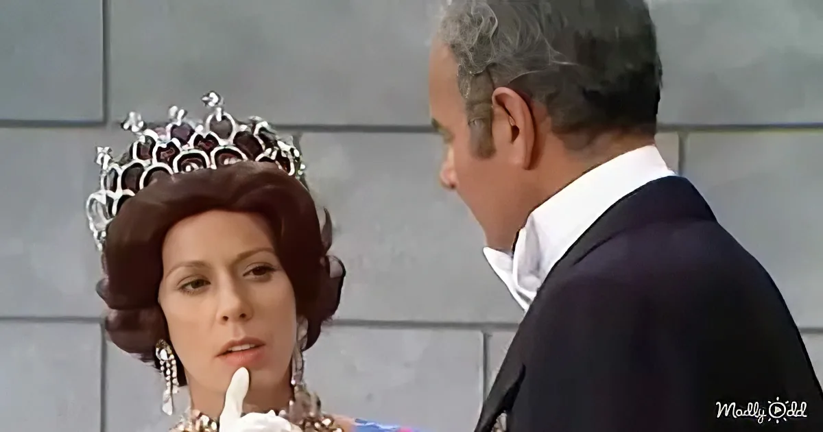 A woman in a tiara and elaborate hairstyle holds a finger to her lips, facing a man in a suit.