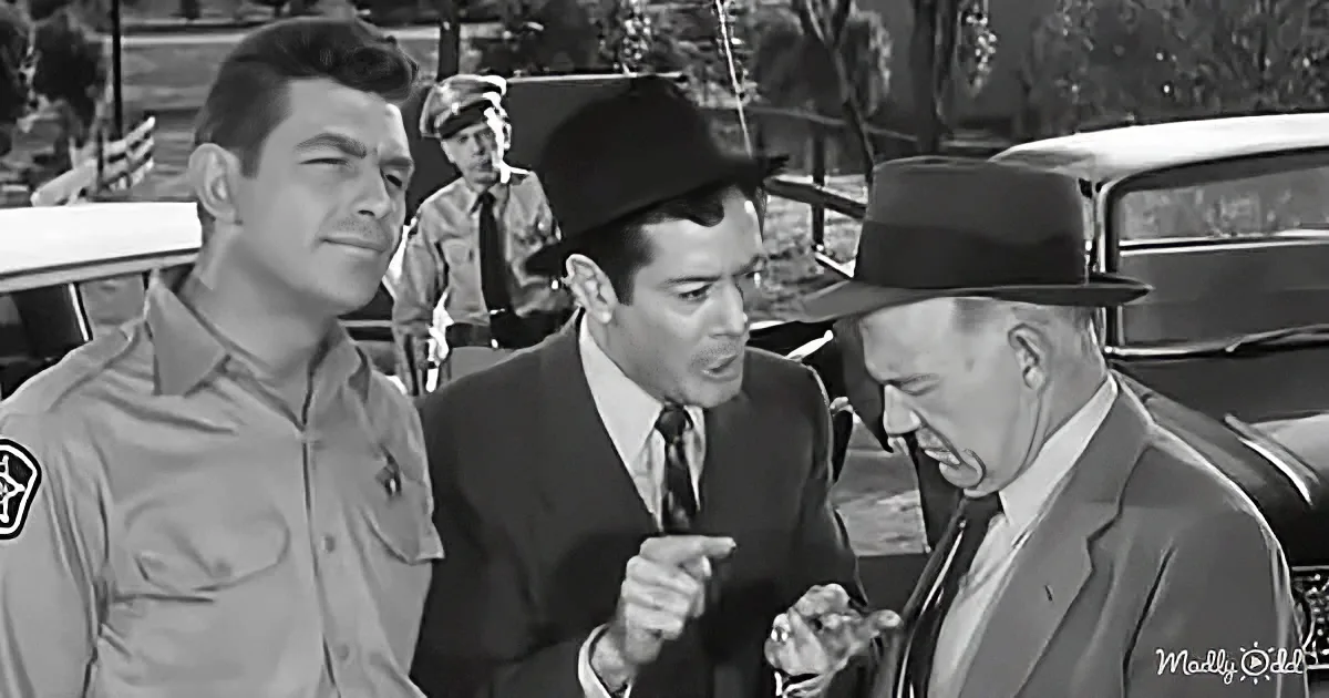 A man in a suit and hat is pointing at another man in a suit and hat, while a uniformed officer looks on.