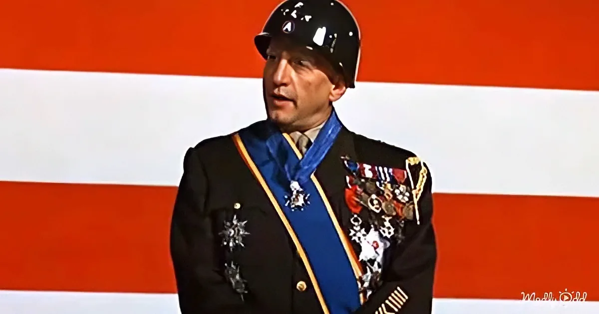 A man in a military uniform with medals and a blue sash stands in front of a red and white background.