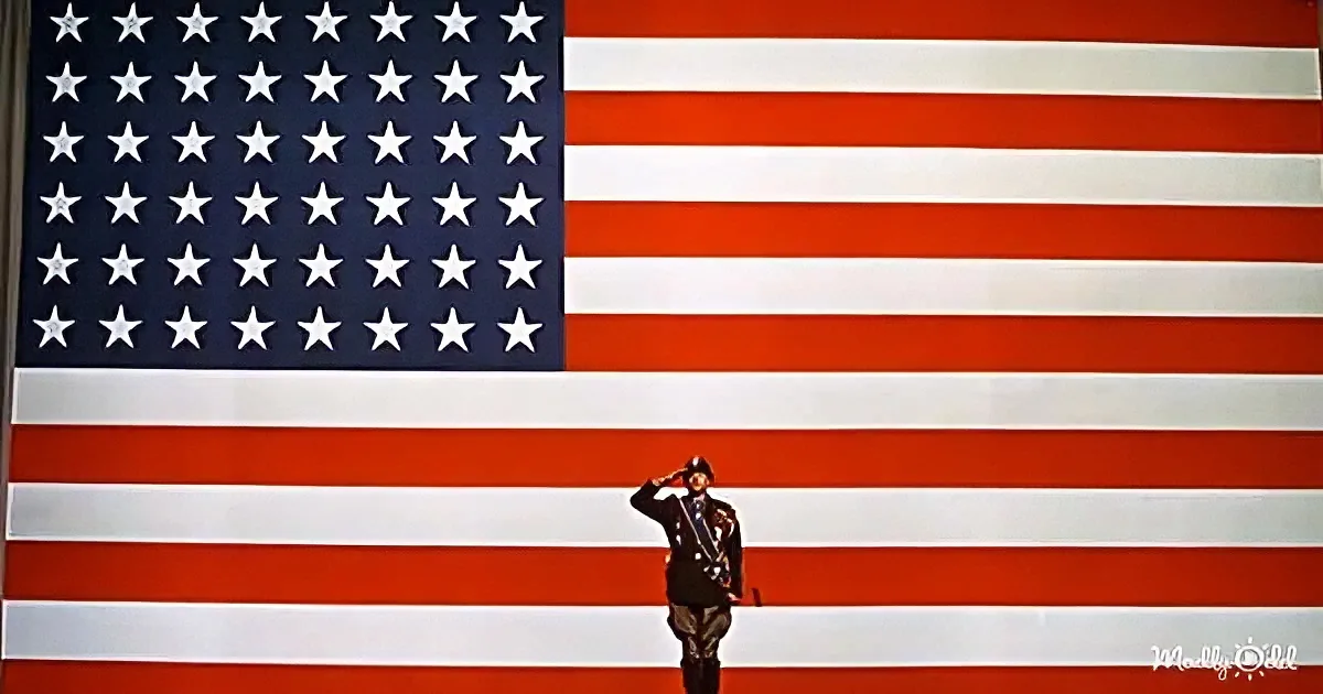 A soldier salutes in front of a large American flag with stars and stripes.