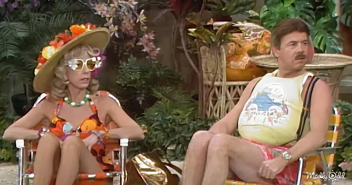 A woman in a colorful bikini and hat sits next to a man in a yellow shirt and red shorts in a tropical setting.