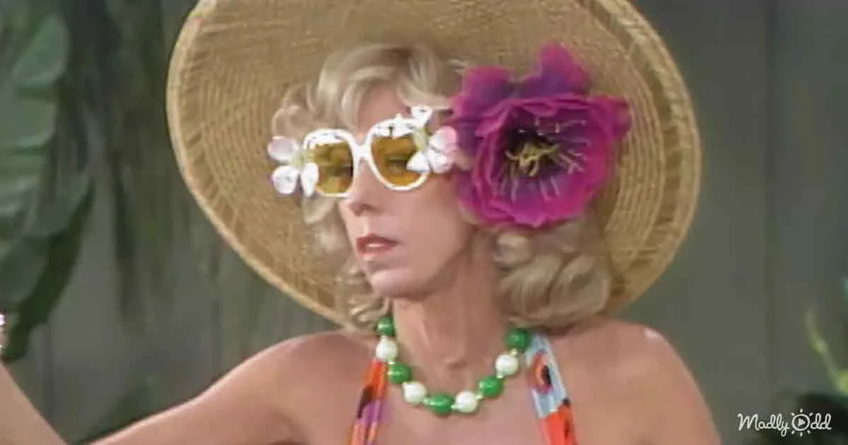 A woman wears a straw hat, sunglasses with flowers, and a colorful necklace, set against a green background.