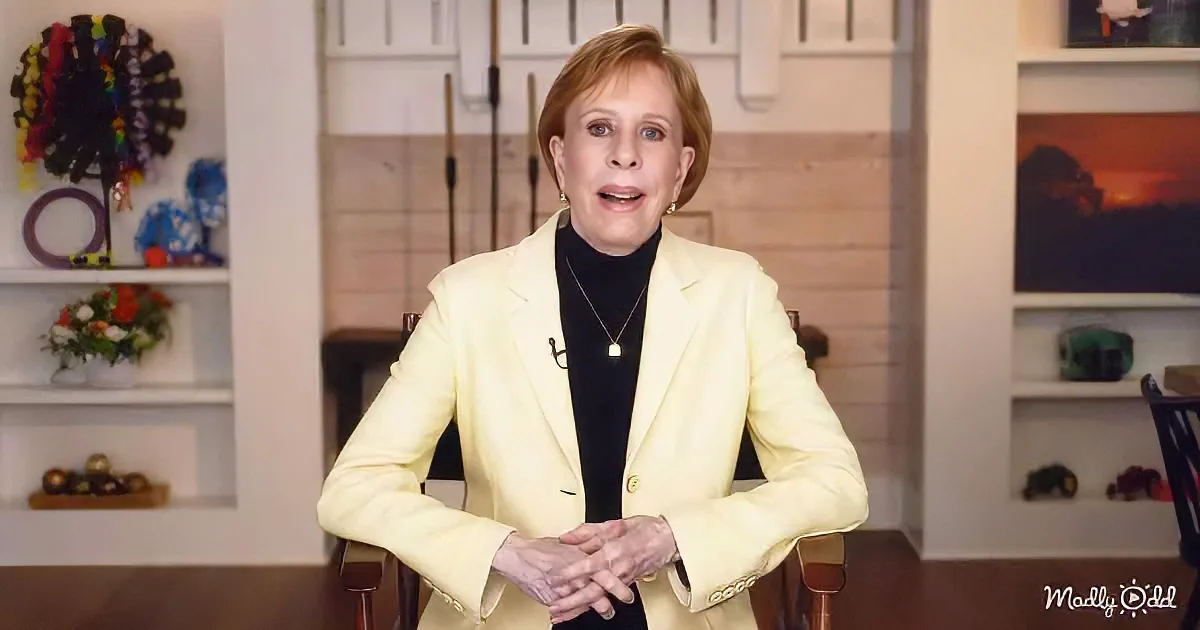 A woman in a yellow blazer sits in a chair, hands clasped, in a room with shelves and decorations.