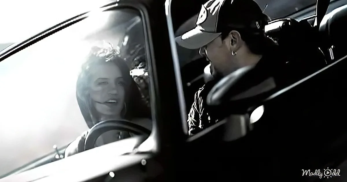 A man in a car, wearing a cap and jacket, looks out the window as a woman smiles in the reflection.