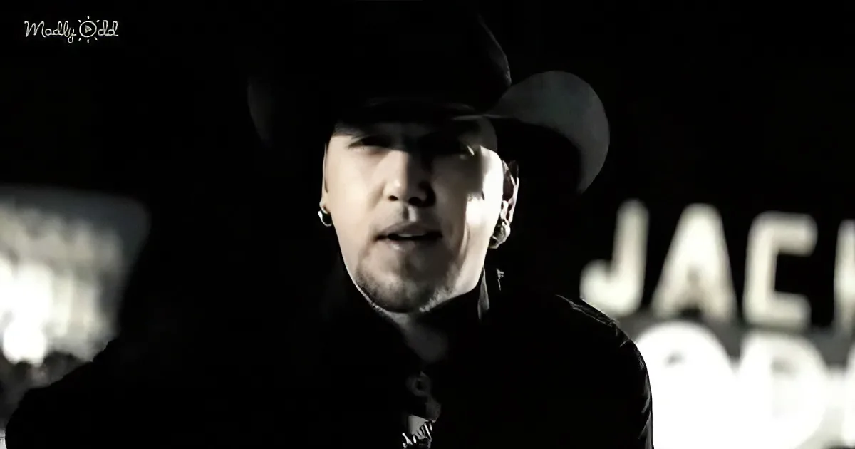 A person wearing a cowboy hat and dark clothing, with a microphone, stands in front of a blurred background with the word...