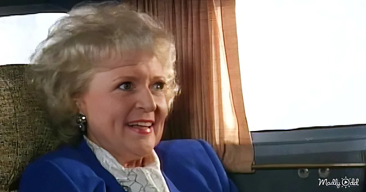 An older woman with curly gray hair, wearing a blue jacket and white shirt, sits in a car with a beige interior and a...