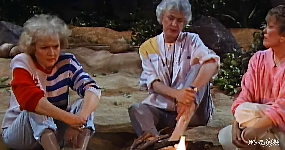 Three women sit outdoors, one in a striped shirt, another in white, and the third in pink, around a fire.