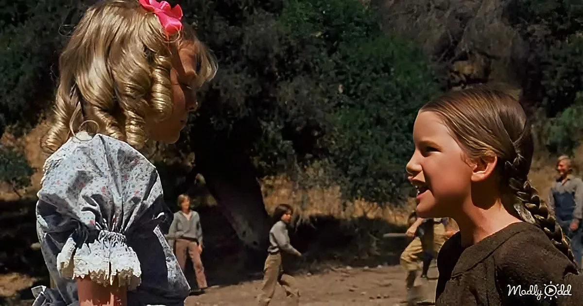 A young girl with braided hair smiles at another girl with a pink bow in a forested area.