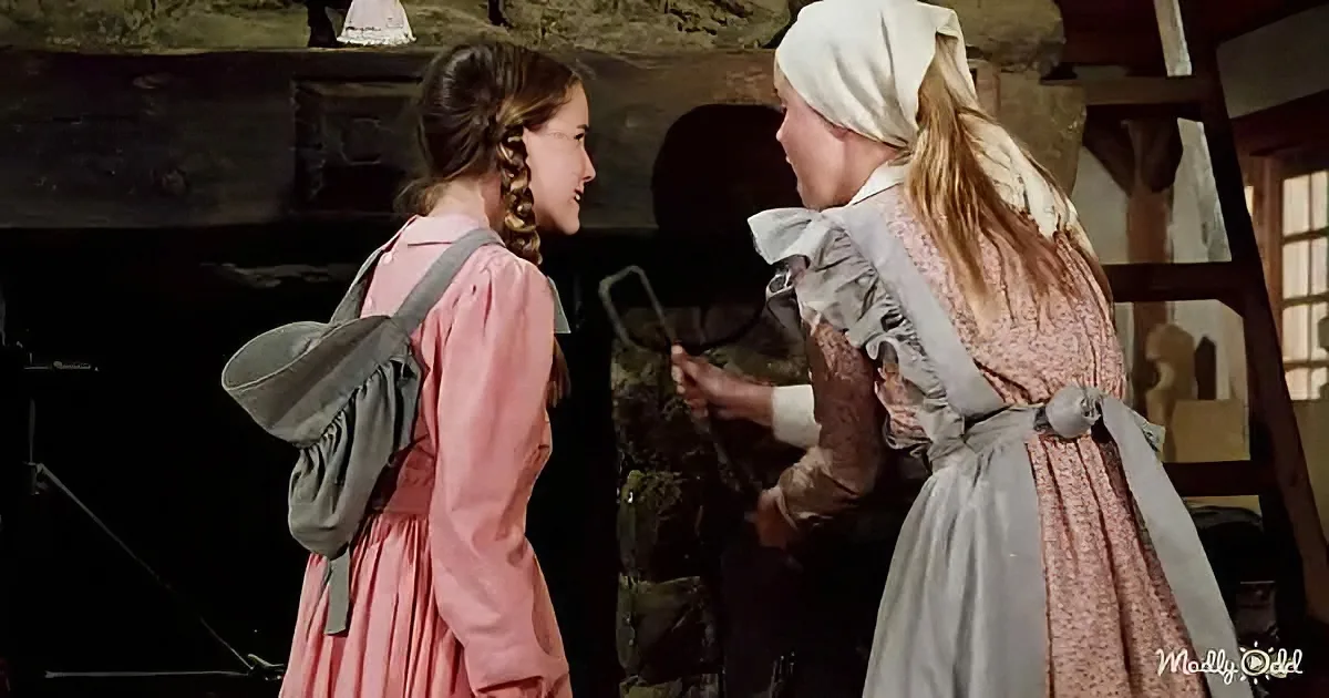 A young girl in a pink dress smiles at another woman in a rustic setting, both wearing period costumes.