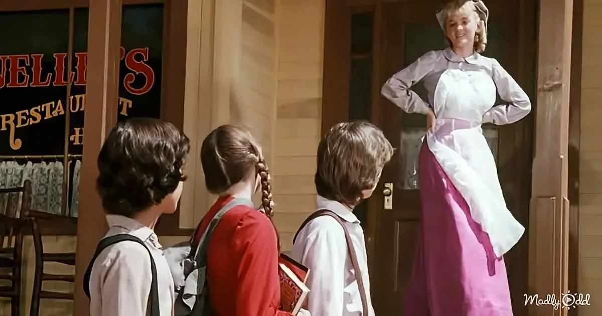 A woman in a pink apron stands in a doorway, looking at three children with backpacks.