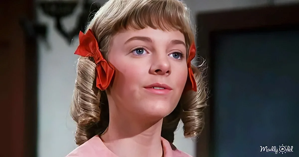 A young girl with blonde hair styled in curls, wearing a red bow, and a pink shirt.