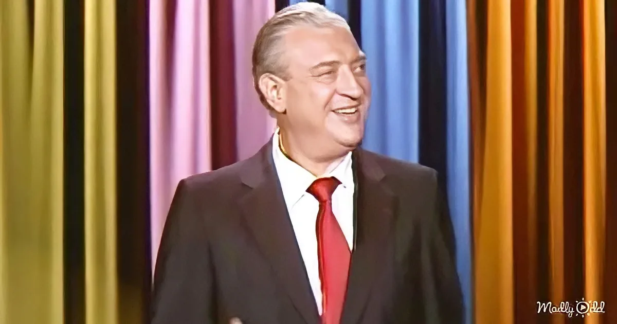 A man in a black suit and red tie smiles against a colorful curtain backdrop.
