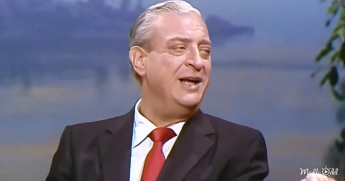 A man in a suit and red tie is speaking, with a blurred background of water and trees.