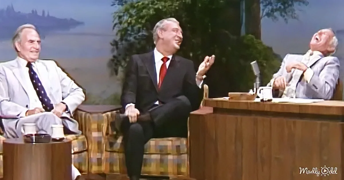 Three men in suits sit on a talk show set, one gesturing animatedly, another laughing, and the third appearing amused.