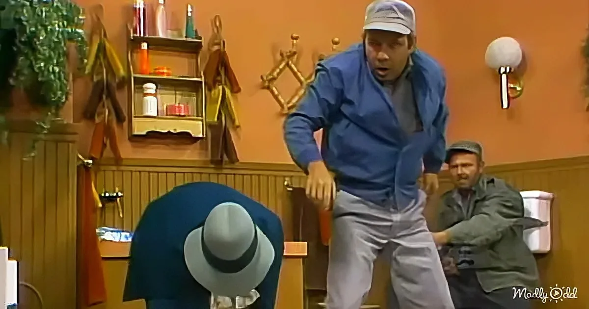 A man in a blue shirt and cap is mid-dance move, surrounded by others in a room with wooden paneling and shelves.
