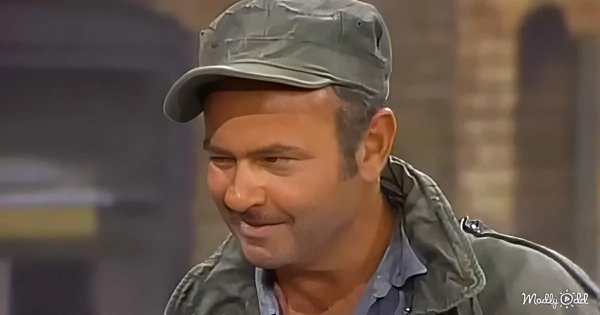 A man wearing a green cap and jacket smiles while sitting, with a blurred background.