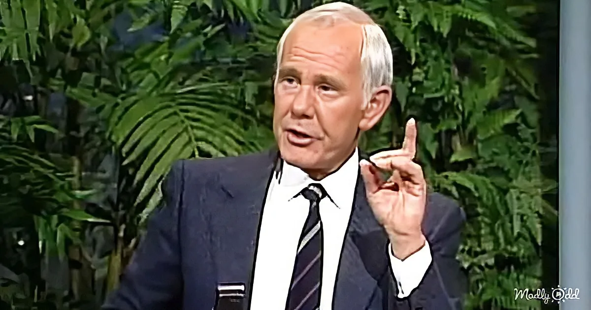 A man in a suit gestures with his right hand while speaking, with green foliage in the background.