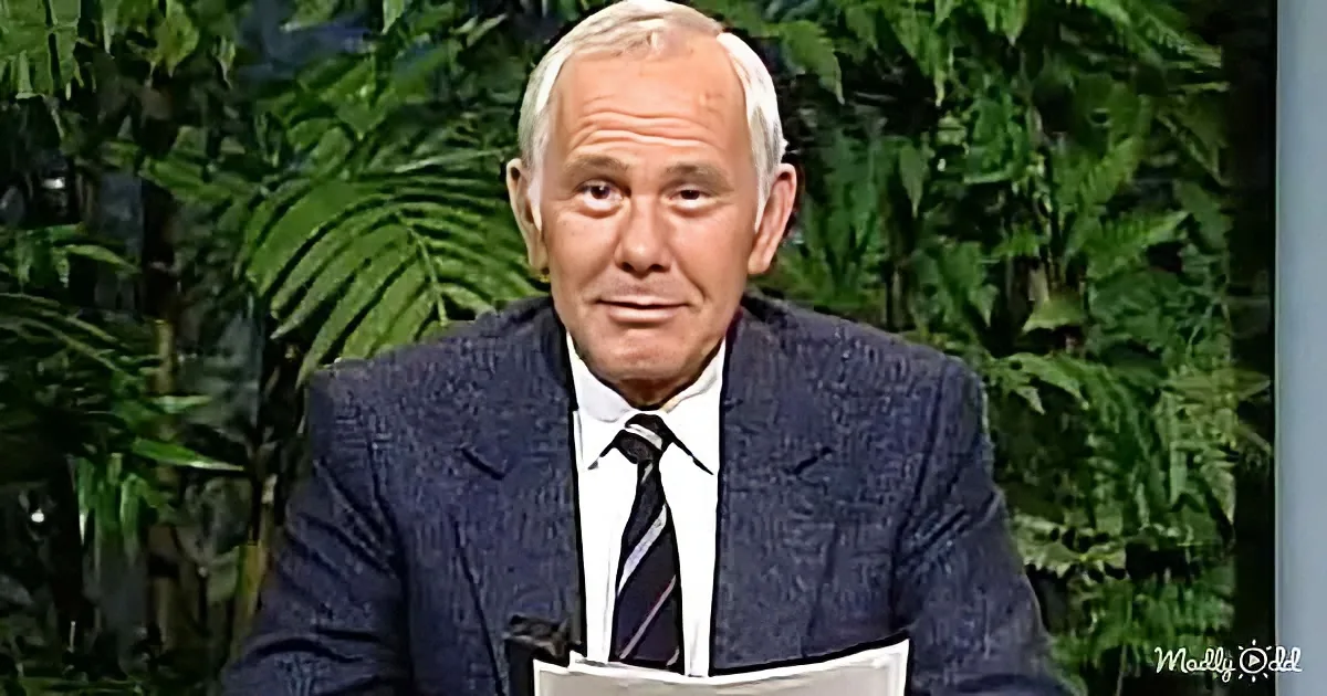 A man in a suit and tie sits at a desk with papers, against a backdrop of green foliage.