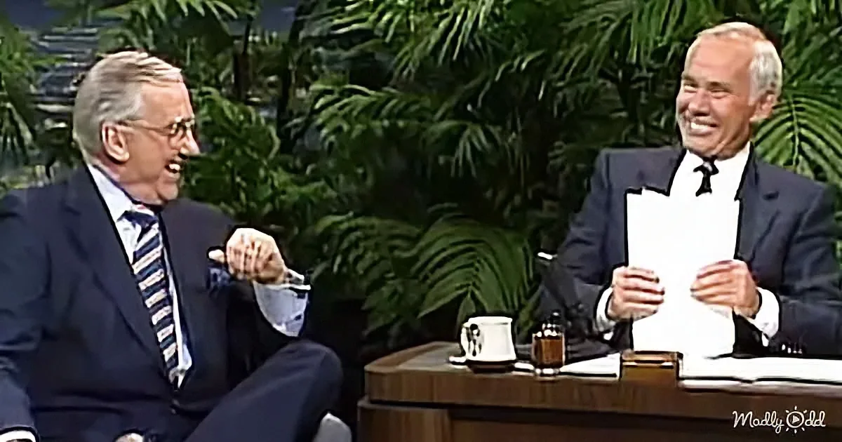 Two men in suits laugh on a talk show set with a desk and plants in the background.