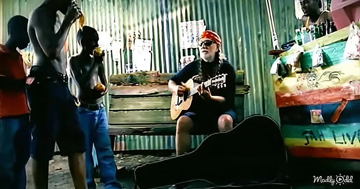 A man in a red bandana plays guitar while three others watch in a rustic setting.