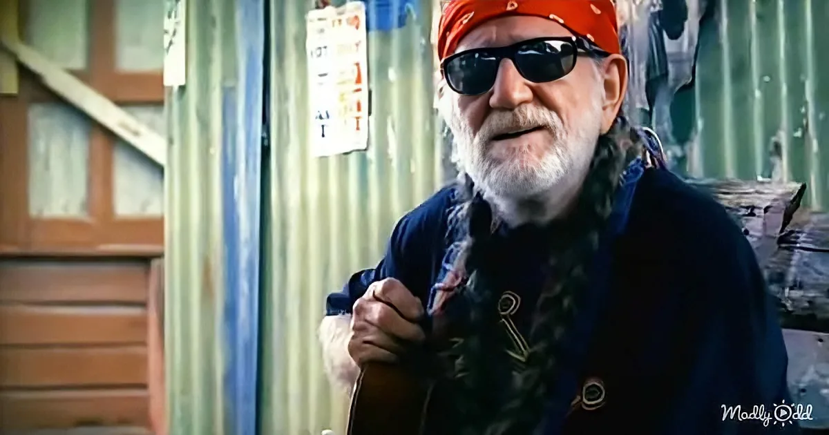 An older man with a white beard and sunglasses, wearing a red bandana, holds a guitar in front of a corrugated metal wall.