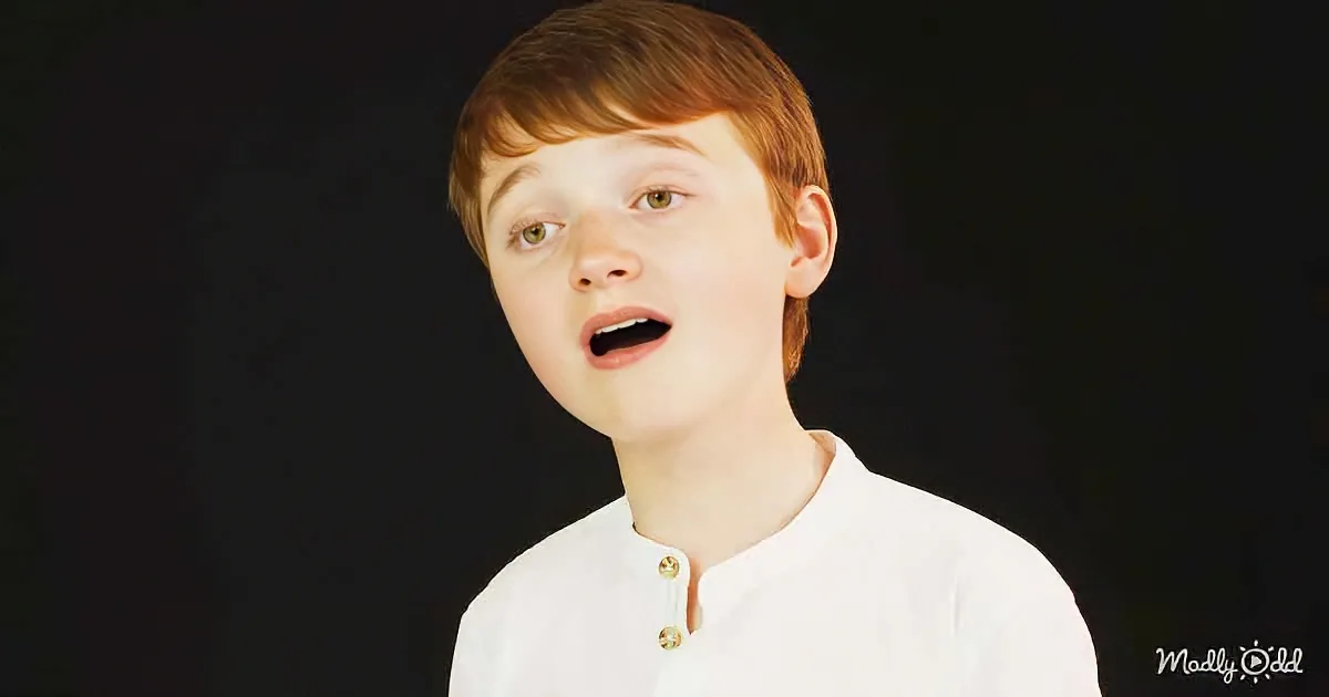 A young boy with red hair and a white shirt stands against a dark background, mouth slightly open.