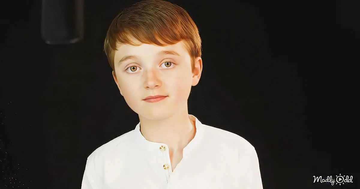 A young boy with short brown hair and a white shirt stands against a dark background.