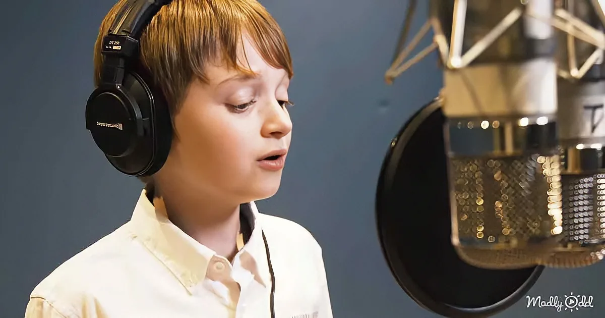 A young boy wearing headphones stands in front of a microphone, seemingly singing or speaking.