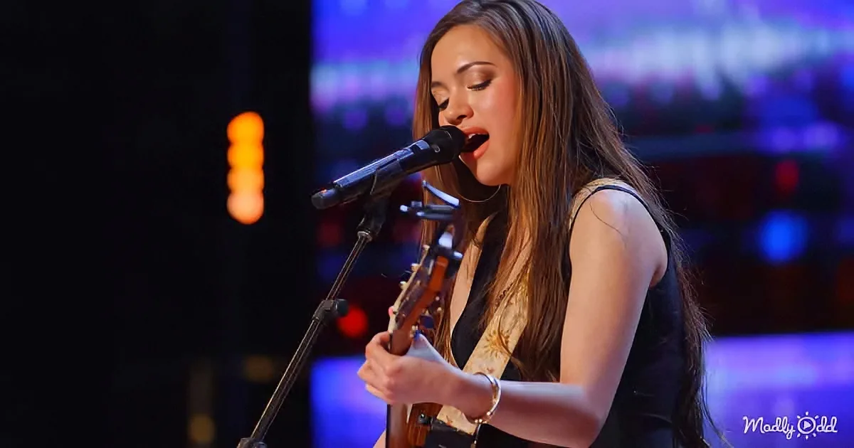 A woman sings passionately into a microphone while holding a guitar on stage.