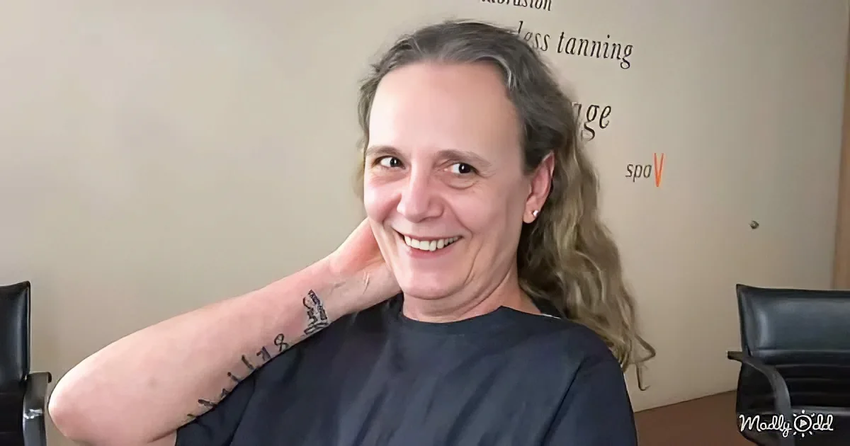 A smiling person with long hair and a tattoo on their arm, wearing a black shirt, sits in a chair.