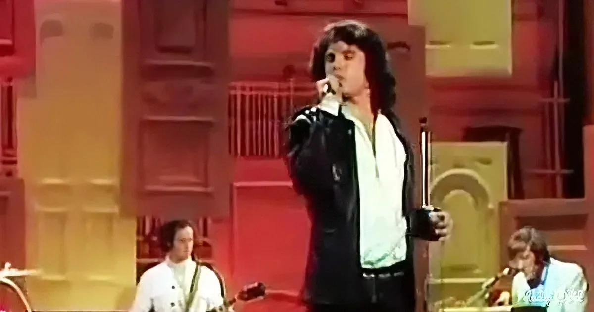 A man in a black leather jacket and white shirt sings into a microphone on stage.