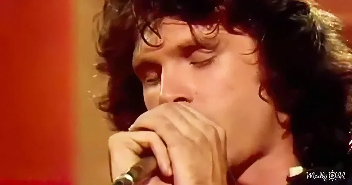 A person with curly hair and glasses holds a microphone close to their mouth, seemingly singing or speaking.