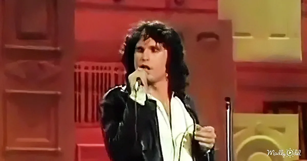 A person with long hair, wearing a black jacket and white shirt, sings into a microphone on stage.