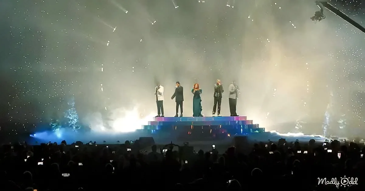 A group of five performers stands on a colorful, illuminated stage with a large audience in front, under dramatic lighting.