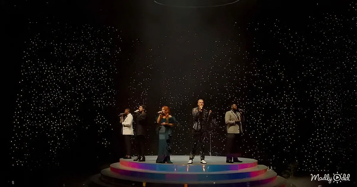 Six performers sing on a circular stage with a starry backdrop.