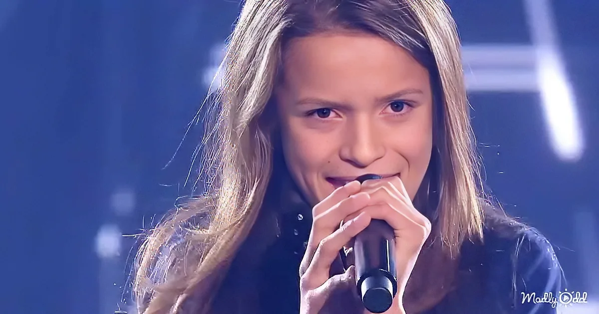 A person with long hair holds a microphone, singing on stage with a dark background.