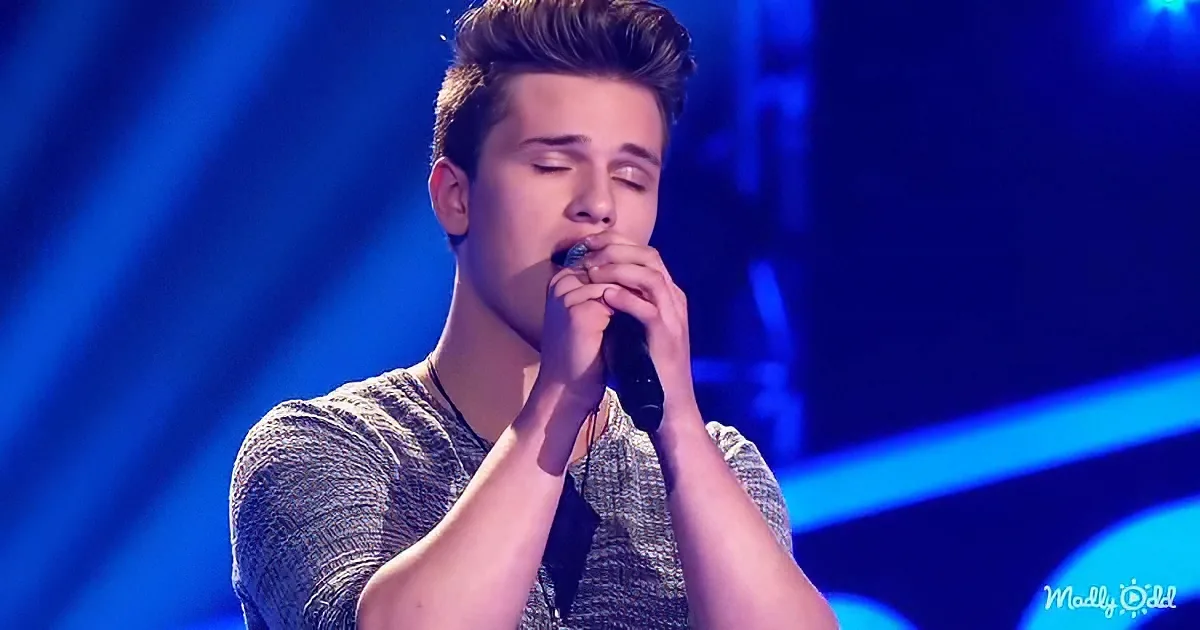 A young man with short hair, wearing a gray shirt, sings passionately into a microphone on stage with blue lighting.
