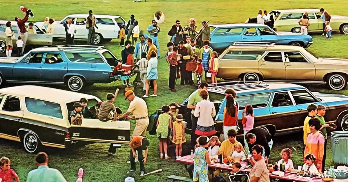 A large group of people, including families and children, gather around vintage station wagons in a grassy field.