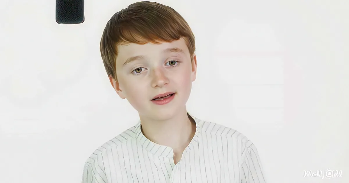 A young boy with short brown hair and green eyes, wearing a white striped shirt, looks slightly to the side with a...