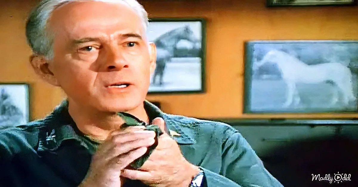 An older man in a green shirt adjusts his collar, with framed horse images on the wall behind him.