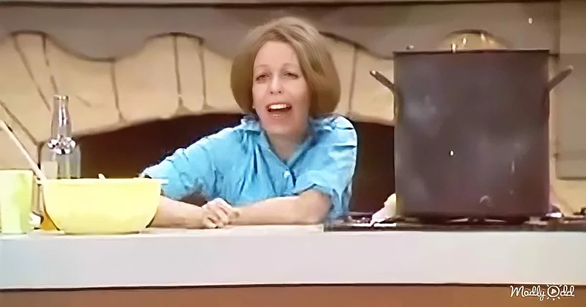 A woman in a blue shirt is smiling at a kitchen counter with a large pot and a yellow bowl.