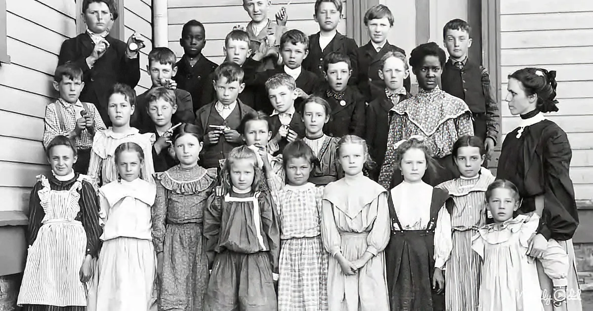 A vintage school group photo with children in period attire, posing in front of a wooden building.
