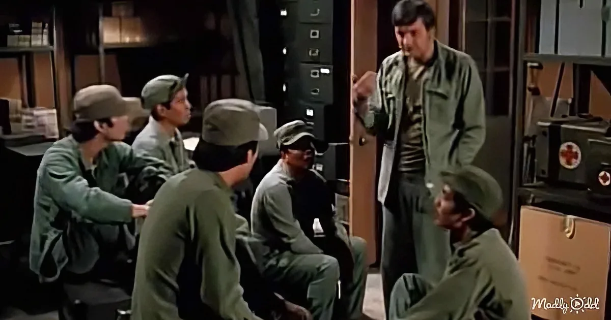 A man in a green uniform stands addressing a group of seated soldiers in a room filled with equipment.