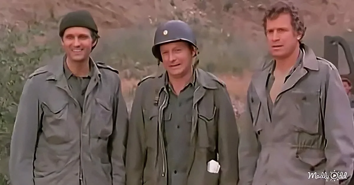 Three men in military uniforms stand outdoors, one wearing a helmet, against a backdrop of hills and vegetation.